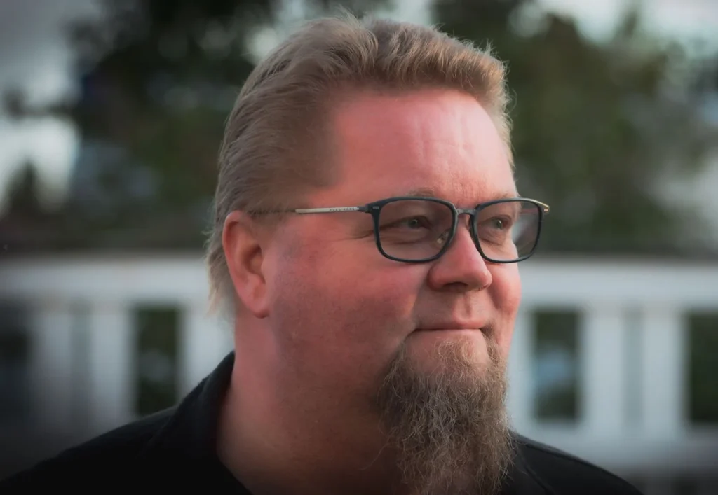 Jarmo Kamsula silmälasit päässä ulkona, taustalla puita ja valkoinen kaide